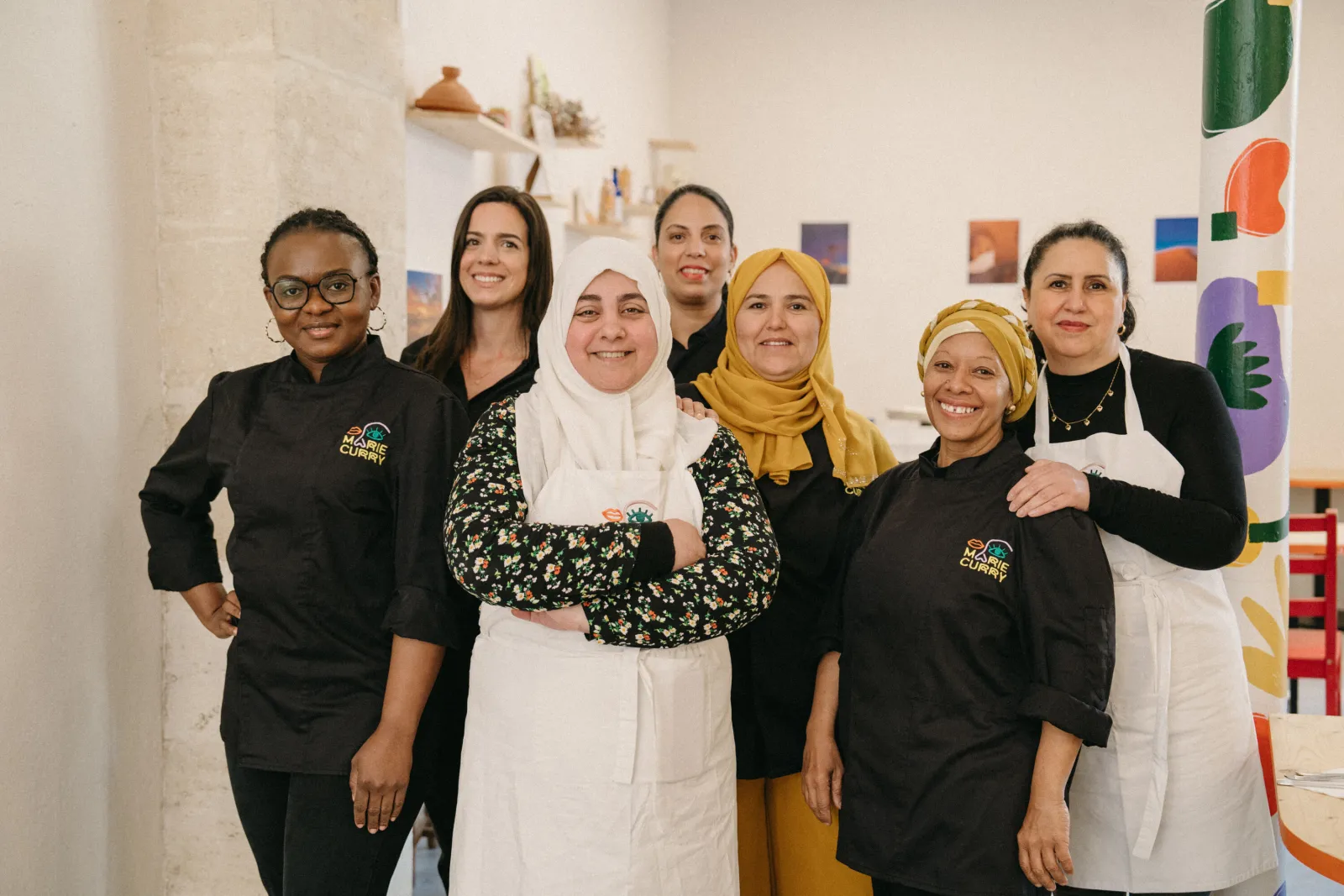 Team of Marie Curry restaurant in Bordeaux: seven smiling women posing together in a white space decorated with shelves and colorful artwork, some wearing the restaurant's black uniform.