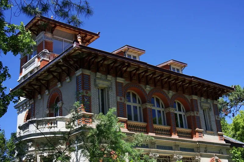 Belle Époque villa in Arcachon's Winter Town featuring red brick façade, arched windows, ornamental balustrades, and pitched roofs surrounded by vegetation.