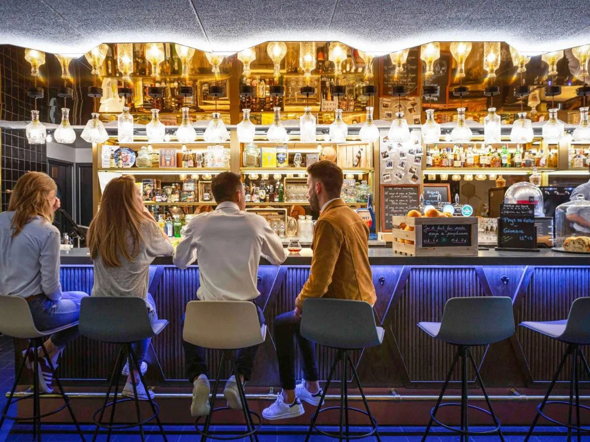 Four customers sitting at the counter of a modern bar with blue lighting, facing a golden mirror and bottles of spirits, at Mercure Hotel Bordeaux Centre.