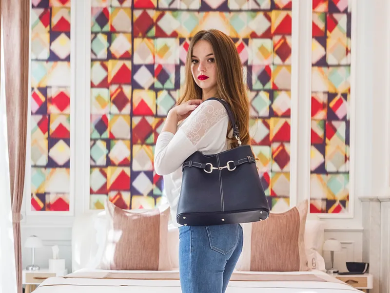 Young woman wearing a black Massip Maroquinerie handbag, dressed in a white blouse and blue jeans, posing in front of a window with colorful stained glass in Bordeaux.