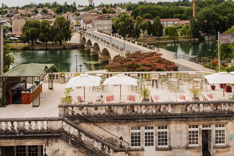 Rooftop terrace with white umbrellas and striped deck chairs overlooking a historic arched bridge spanning a river, with buildings and cityscape beyond