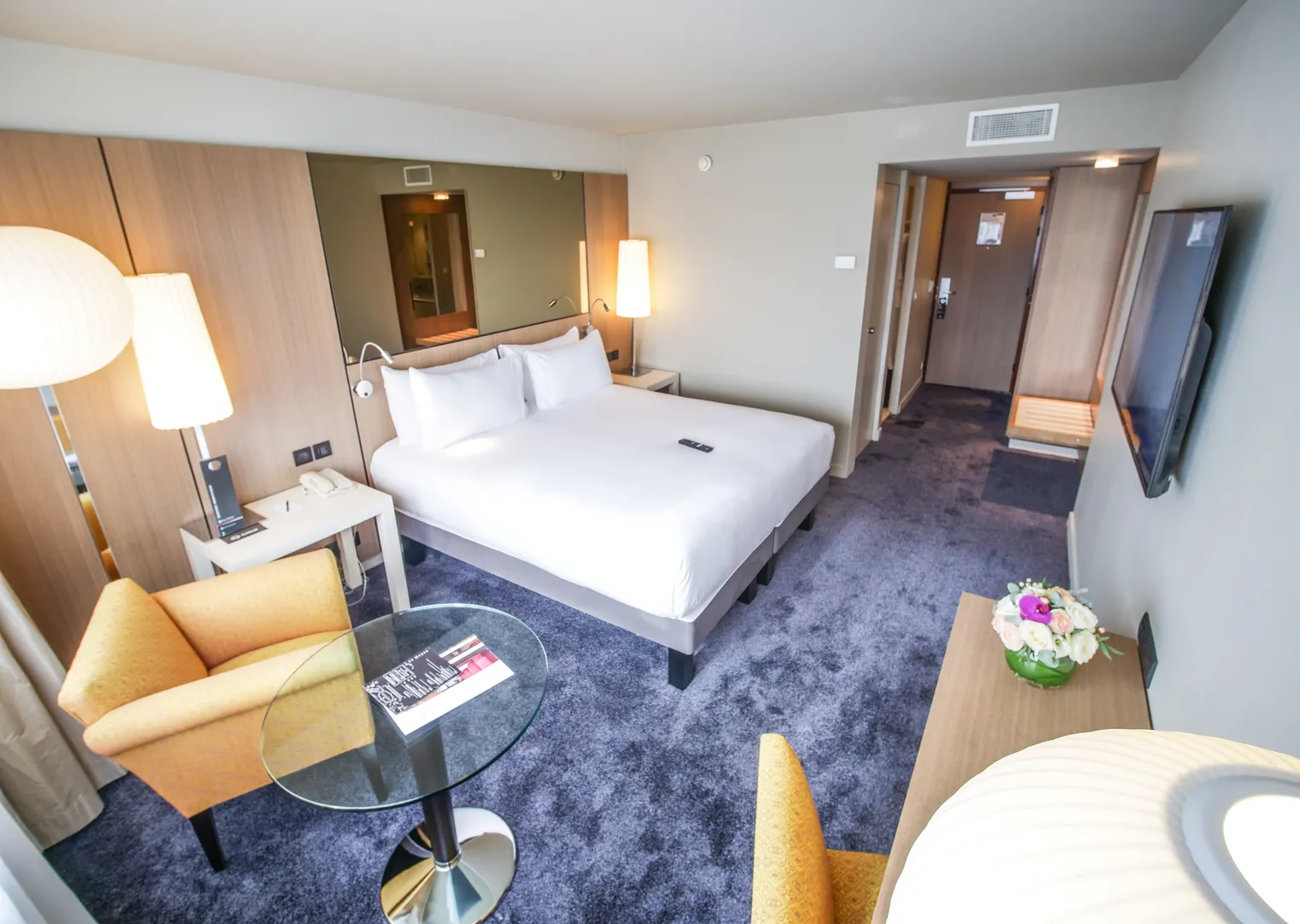 Modern hotel room at Pullman Bordeaux Lac featuring a king-size bed, yellow armchair, work desk, bathroom and entrance view