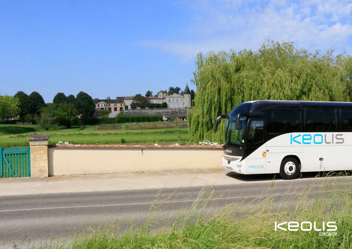 A white and black Keolis Gironde coach bus parked on the roadside in Gironde, with a château and green vegetation in the background.