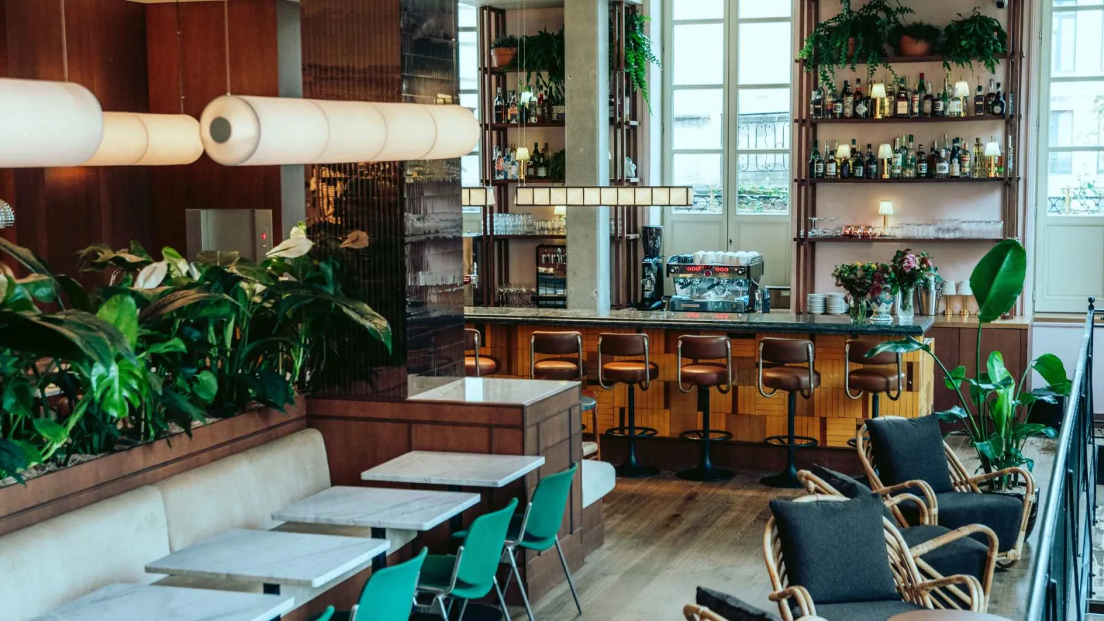 Modern bar-restaurant interior in Bordeaux featuring wooden counter, bar stools, green plants, liquor bottle shelves, elegant furnishings and large windows with views.