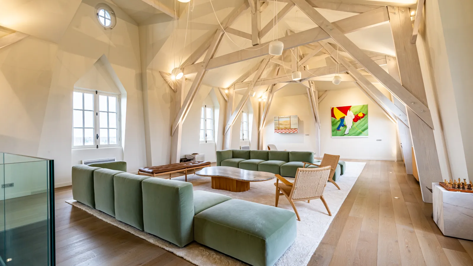 Spacious loft interior with white exposed beams, green sofas, vintage wooden furniture, large windows, and colorful artwork on the walls.