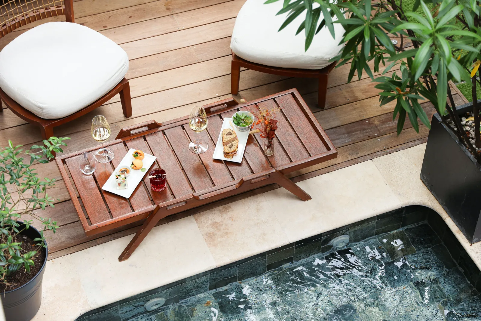 Elegant terrace at a Bordeaux boutique hotel featuring a wooden coffee table with seafood appetizers, white wine glasses, white cushioned chairs, and an adjacent swimming pool surrounded by greenery.