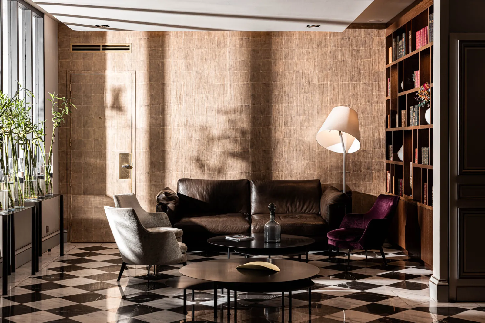 Elegant living room at Hôtel Villas Foch in Bordeaux featuring a black leather sofa, gray and burgundy armchairs, geometric black and white flooring, beige travertine walls, light wood shelving, and a white pendant light.
