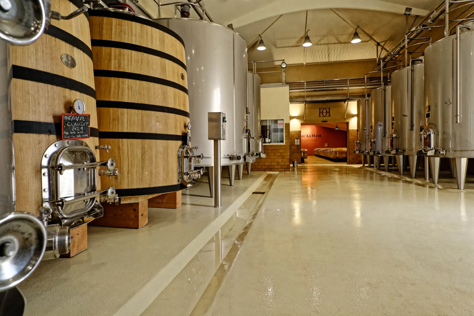 Interior of Château La Haye winery in Bordeaux featuring oak barrels with black bands, stainless steel tanks, and polished concrete floor