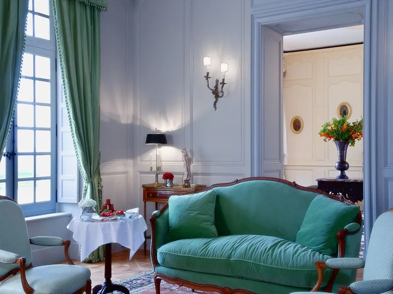 Elegant living room in Château Soutard, Bordeaux, featuring a green emerald sofa, gray armchairs, a window with green curtains, a white side table, a golden wall sconce, and a colorful flower arrangement.