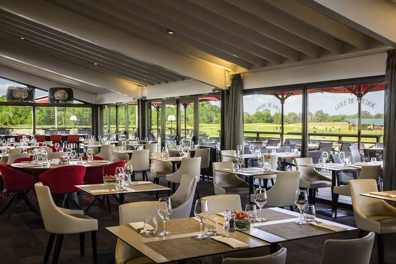 Upscale dining room with floor-to-ceiling windows overlooking a golf course, featuring elegant tables with cream and burgundy chairs, exposed beam ceiling, refined décor and abundant natural light.