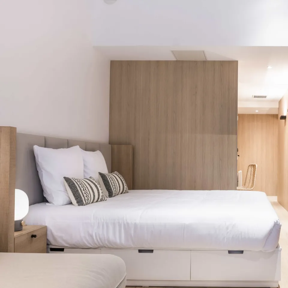 Modern hostel bedroom featuring a double bed with white linens, gray pillows and striped cushions, light wood headboard, white integrated storage and wooden partition wall at Central Hostel Bordeaux