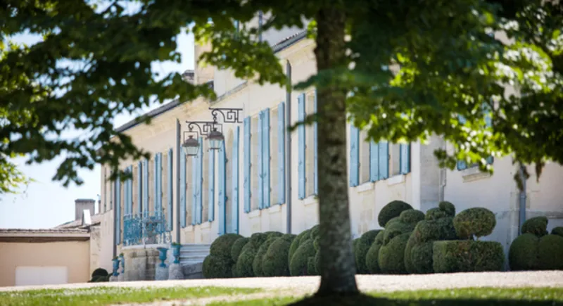 Facade of Château Fourcas Hosten in Bordeaux featuring distinctive blue shutters, surrounded by manicured boxwood hedges and trees under clear sky.