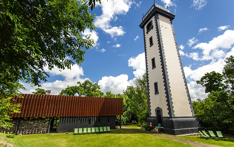 White modern tower with black base on Patiras Island in Bordeaux, accompanied by a building with red roofs, surrounded by green spaces and trees.