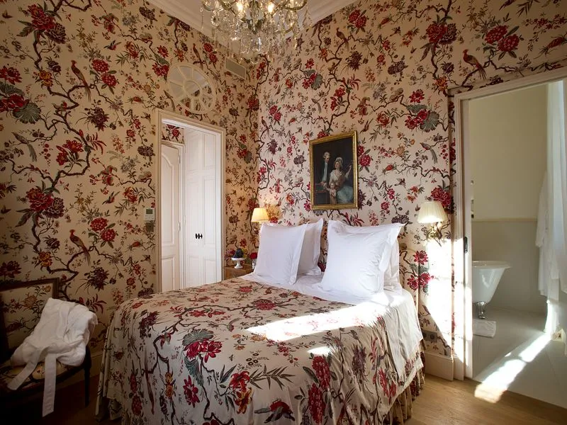 Elegant bedroom at Château Soutard in Bordeaux featuring red and beige floral wallpaper, white bed with matching cushions, crystal chandelier, antique portrait and white doors