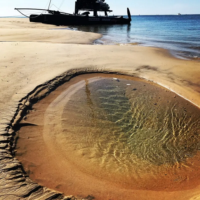 Black boat beached on sandy shore with a circular natural tidal pool in the foreground, water reflections under sunlight, blue sea in the background