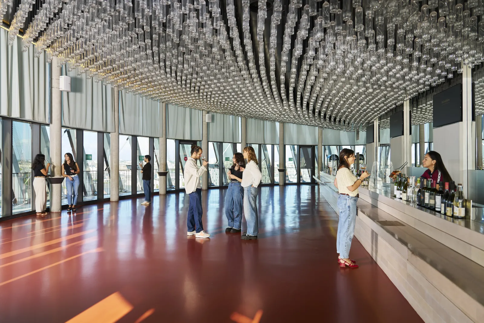 Modern interior of the Cité du Vin in Bordeaux featuring a glass and metal architectural ceiling, red marble flooring, panoramic windows, and visitors exploring the space.