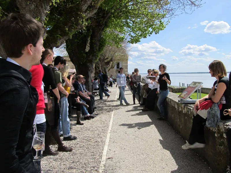 Group of visitors gathered on a paved path along the fortifications of Blaye Citadel, overlooking the Gironde estuary with century-old trees.