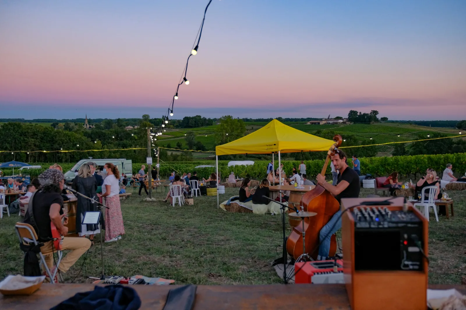 Outdoor concert at sunset in Bordeaux vineyards, with musicians playing cello and wind instruments, audience seated under yellow umbrellas, rural landscape in the background.
