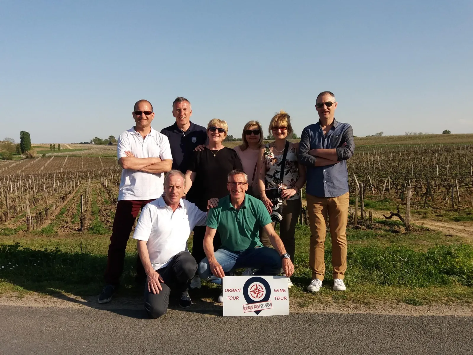 Group of eight people posing in a Bordeaux vineyard with an Urban Wine Tour sign, under clear skies.