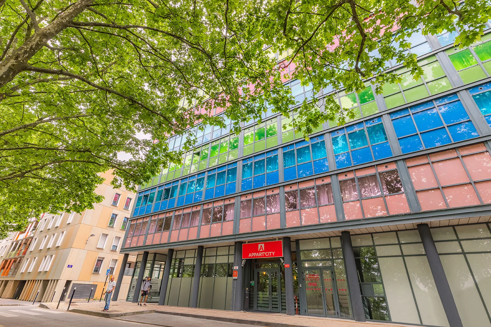 Modern facade of Appart'City Classic in Bordeaux featuring blue and green tinted windows, red ground-floor signage, lush green tree foliage in foreground, historic yellow buildings nearby.