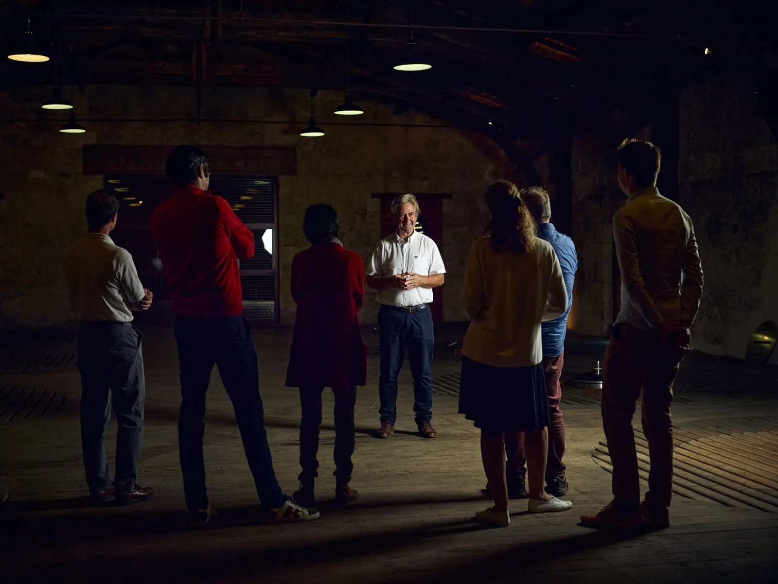 A guide in a white shirt explains to a group of visitors gathered in a circle in an underground cellar of Château Desmirail in Bordeaux, lit by ceiling lamps.
