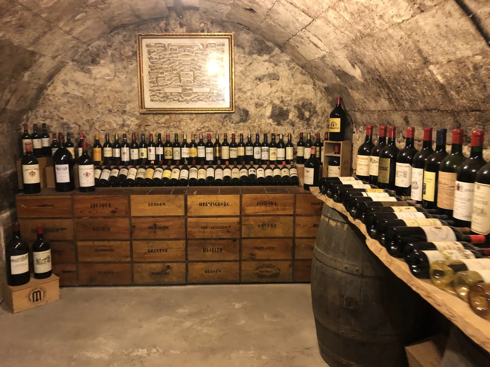 Underground cellar displaying hundreds of wine bottles arranged on wooden shelves, antique wooden crates labeled with wine regions, and a wooden barrel in the foreground, within historic stone architecture.