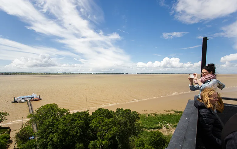 Two people viewing the Gironde estuary from an observation platform at Patiras Island, Bordeaux. Panoramic view of muddy waters and distant shores under partly cloudy skies.