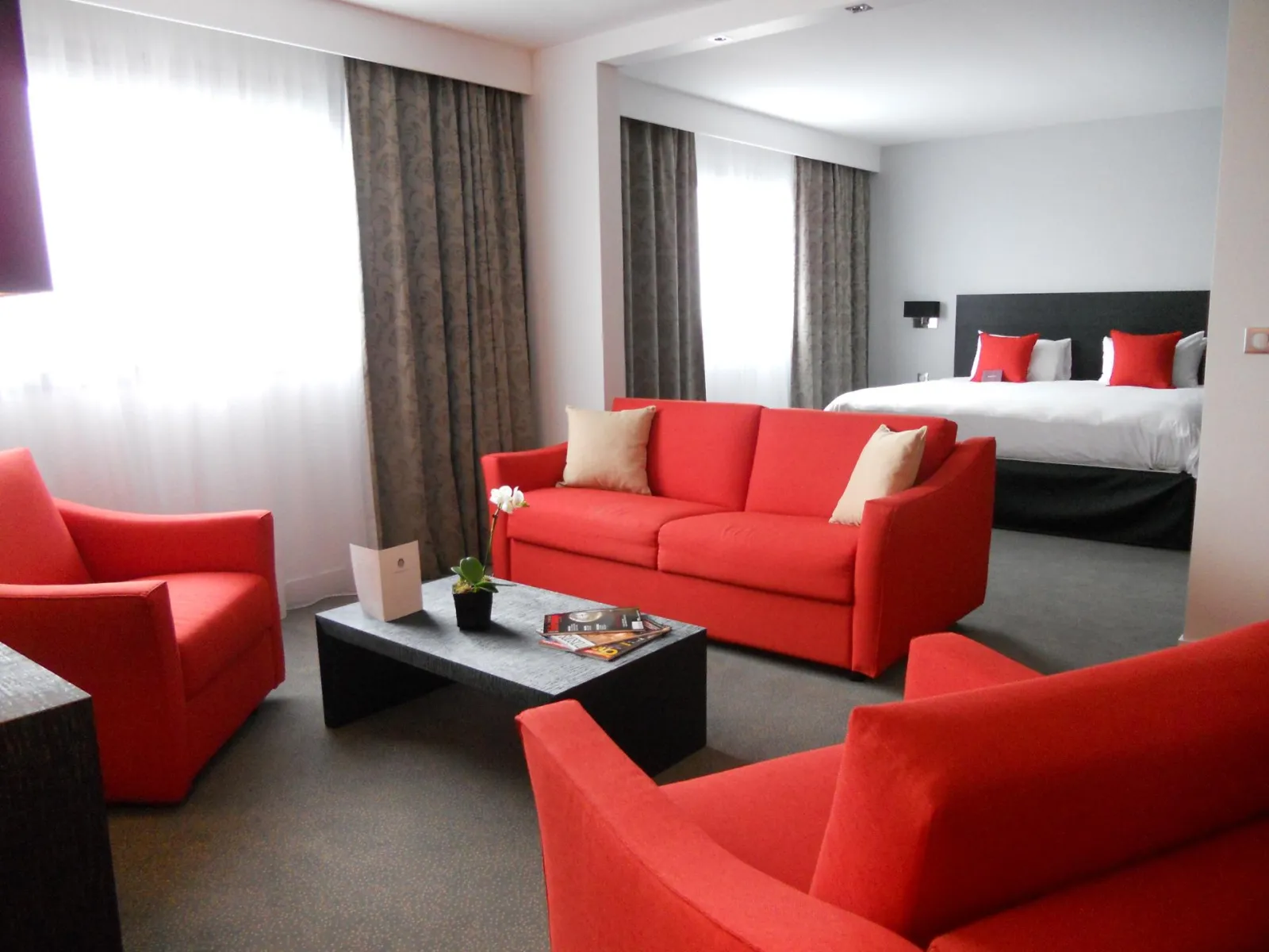 Spacious suite at Mercure Bordeaux-Lac hotel featuring red sofa and matching armchairs, black coffee table, gray curtains, and bed in the background