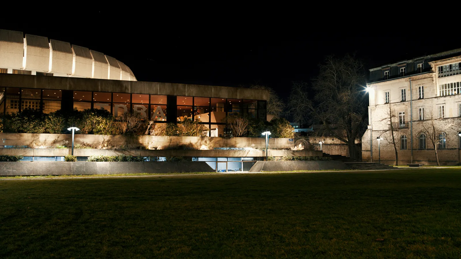 Night view of the National Theater of Bordeaux in Aquitaine, featuring modern illuminated architecture with glass facade and adjacent classical building overlooking a lawn and the Garonne river