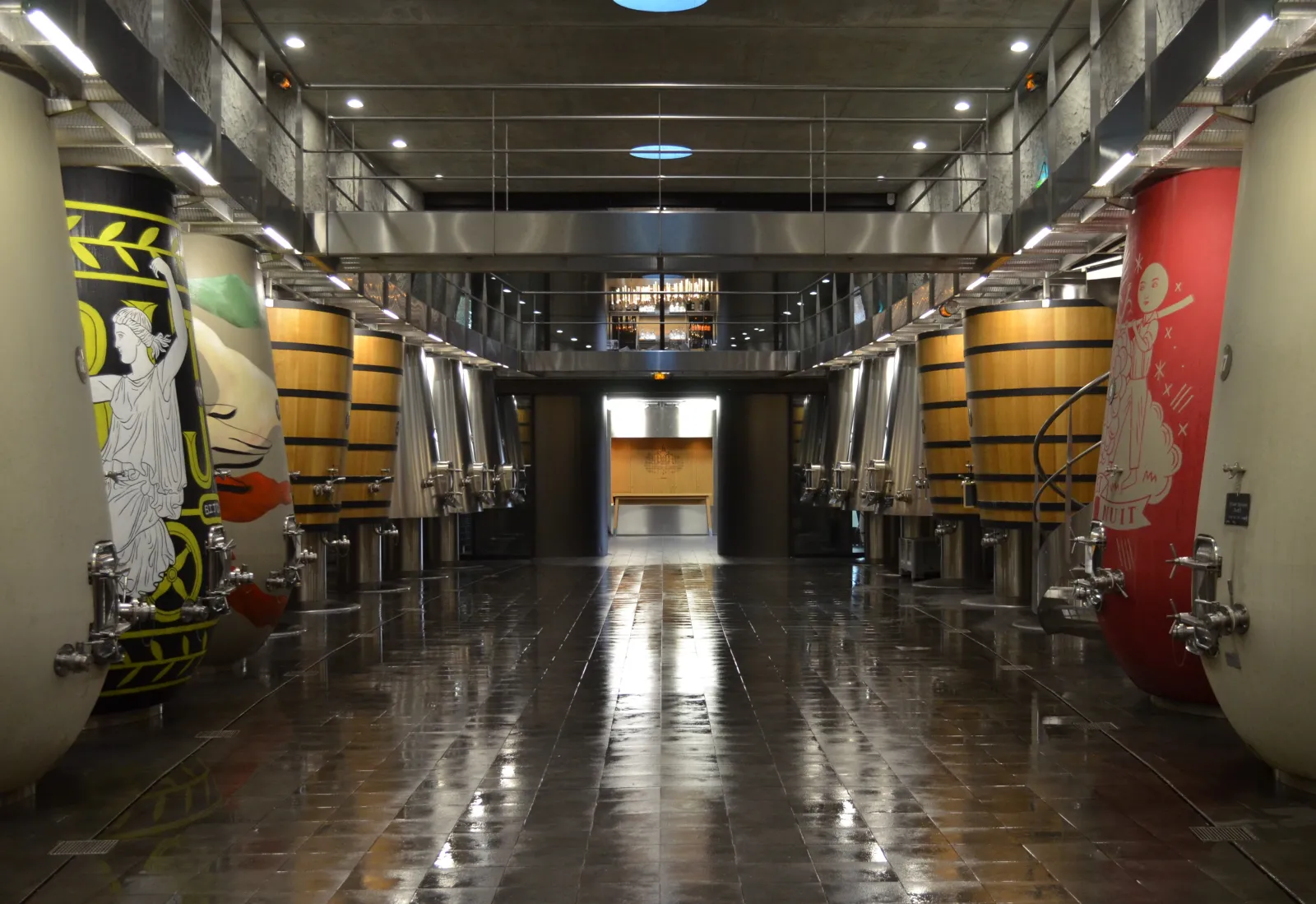 Modern wine cellar interior featuring rows of yellow and black oak barrels, wet reflective floor, artistic wall panels, and metal doors at the end of the corridor.
