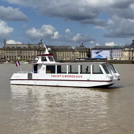 The Yacht of Bordeaux, a white cruise boat sailing on the Garonne River with 18th-century Bordeaux architecture in the background under a cloudy sky.