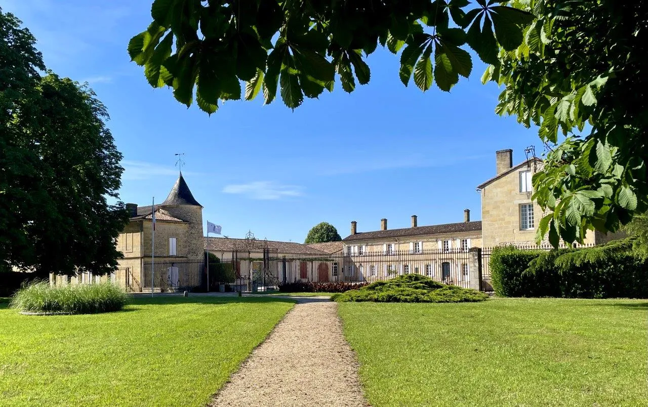 Courtyard of Château de Portets near Bordeaux featuring stone buildings with a conical tower, central pathway, and manicured green lawns against a clear blue sky.