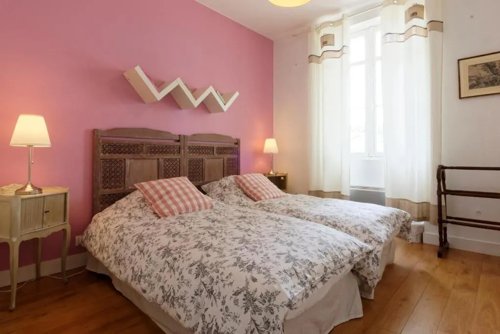 Elegant bedroom featuring a vibrant pink accent wall, wooden carved bed frame, floral patterned bedding with pink and white striped pillows, bedside lamps, large window with white curtains, wooden flooring and classic furniture.