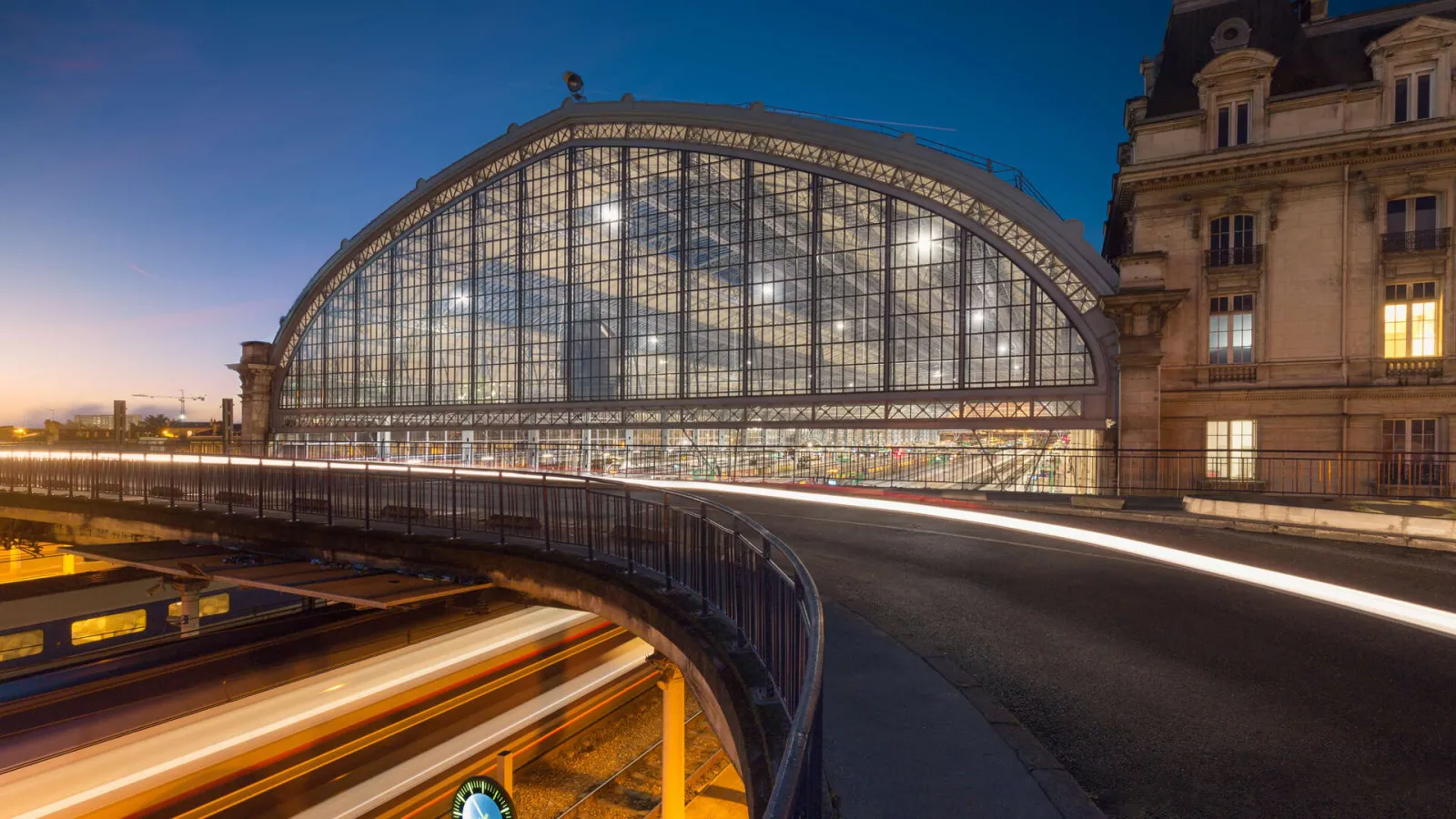 Night view of Bordeaux Saint-Jean railway station featuring its large illuminated metal and glass arch, historic stone façade, and light trails from passing traffic.