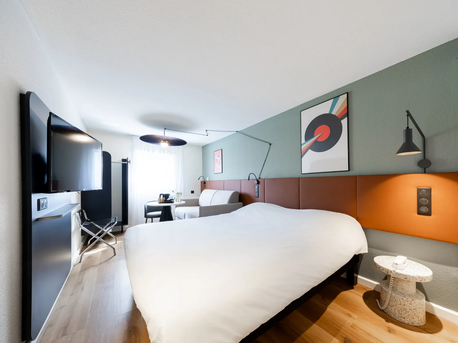 Modern hotel room featuring a large white bed with orange and brown headboard, mint green wall, flat-screen TV, desk, and contemporary decor in Bordeaux