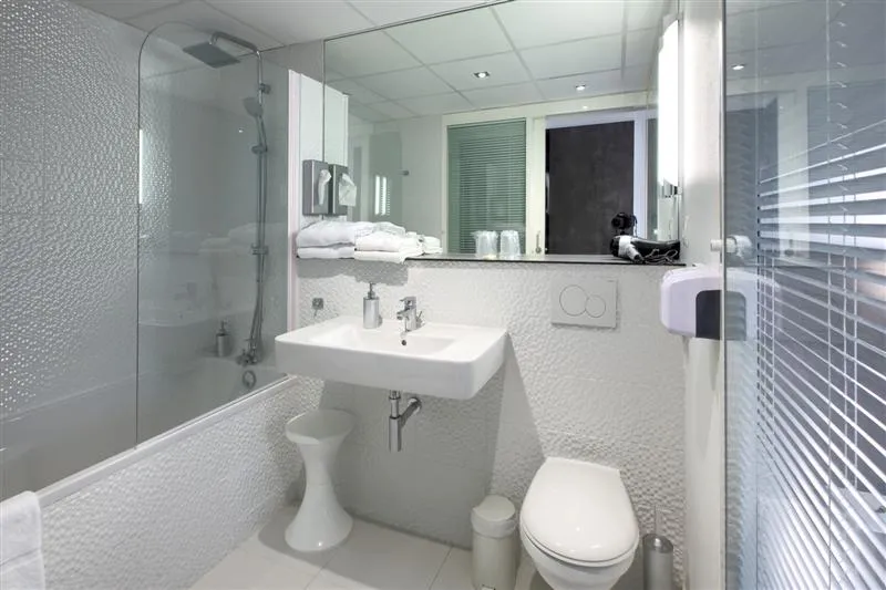Modern and bright bathroom with glass shower enclosure, white sink, toilet, illuminated mirror and bathroom accessories at Au Comté d'Ornon Hotel in Bordeaux