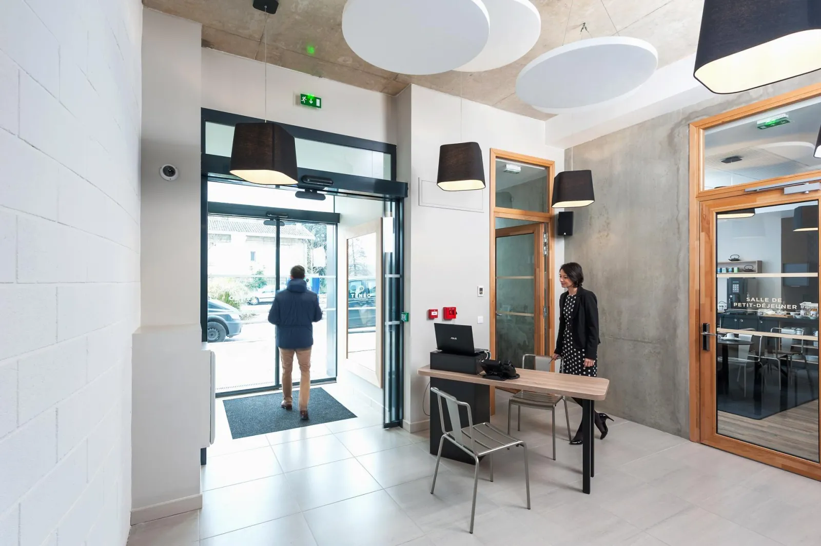 Modern reception area of an aparthotel in Talence, Bordeaux, featuring contemporary design, glass doors, reception desk with staff, and entering guest, with circular ceiling lights.