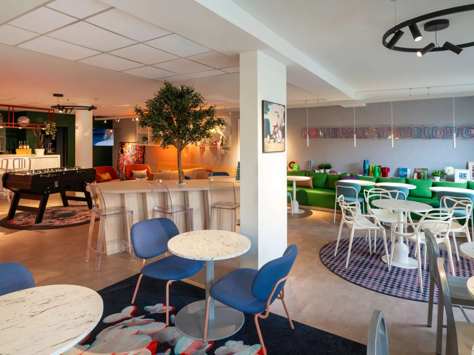Modern common area of the Ibis Styles Bordeaux Centre Gare hotel featuring round tables, blue chairs, bar counter, foosball table and colorful lounge area.