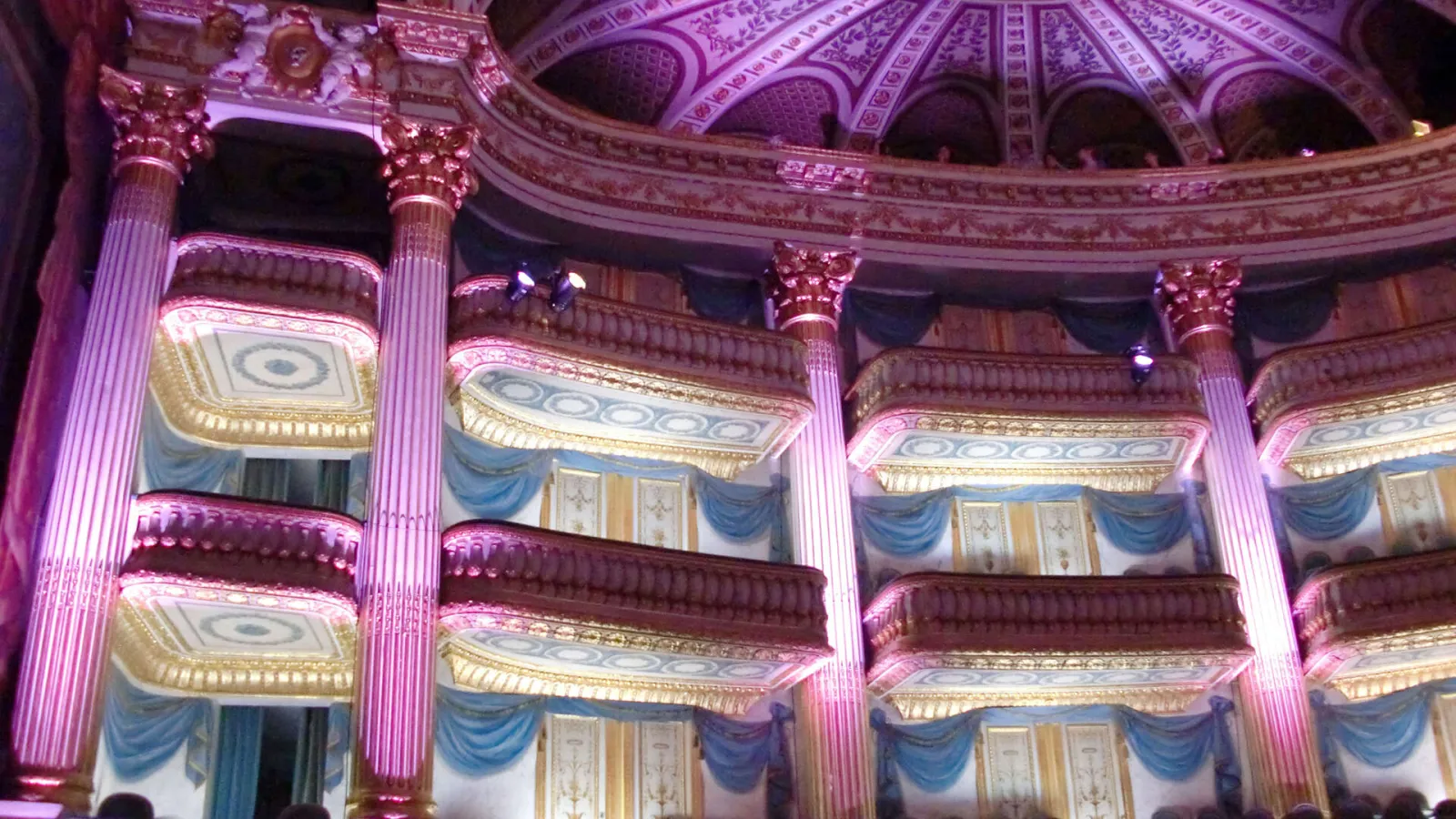 Interior of the Grand Theatre of Bordeaux with pink and purple lighting, featuring ornate balconies, Corinthian columns, blue drapes, and elaborately decorated dome