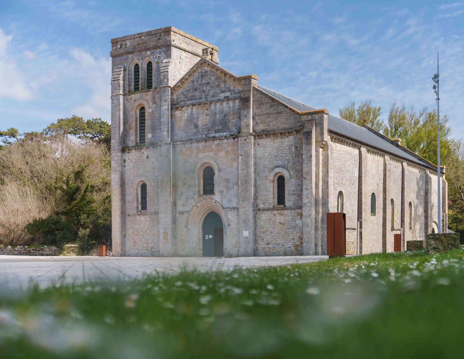 Basilica of Notre-Dame-de-la-fin-des-Terres in Bordeaux, stone church with square tower and Romanesque façade, surrounded by greenery under blue sky.
