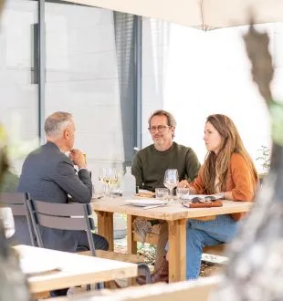 Three people seated at a table in a restaurant at Chais de la Fleur in Bordeaux, conversing over glasses and plates
