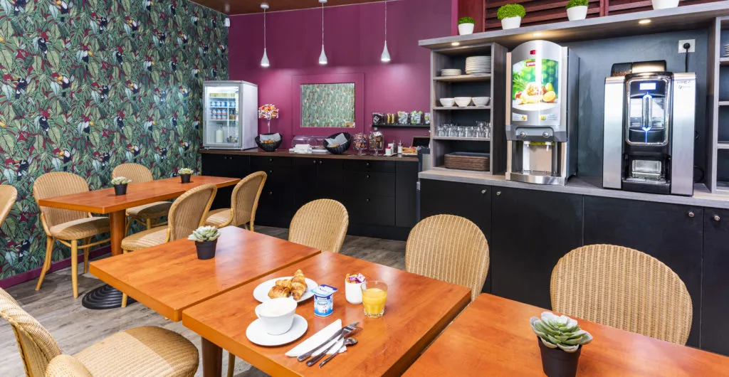 Dining and common kitchen area of an aparthotel featuring wooden tables, rattan chairs, magenta walls with floral wallpaper, and modern appliances including refrigerator, coffee machine, and shelving.