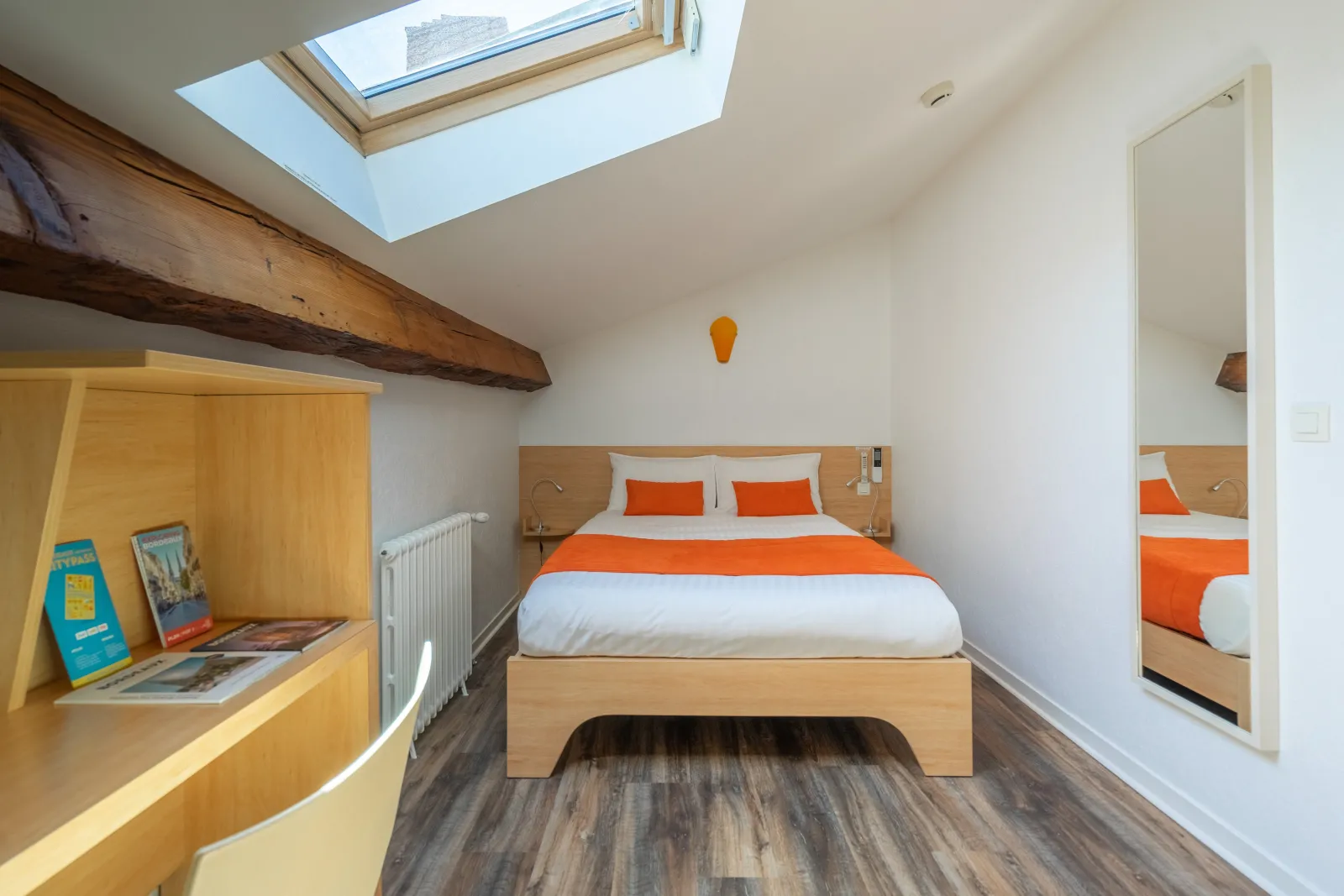 Attic bedroom at Hôtel des Voyageurs in Bordeaux featuring a double bed with orange and white bedding, light wood furniture, exposed beam, mirror, skylight window, and gray parquet flooring.