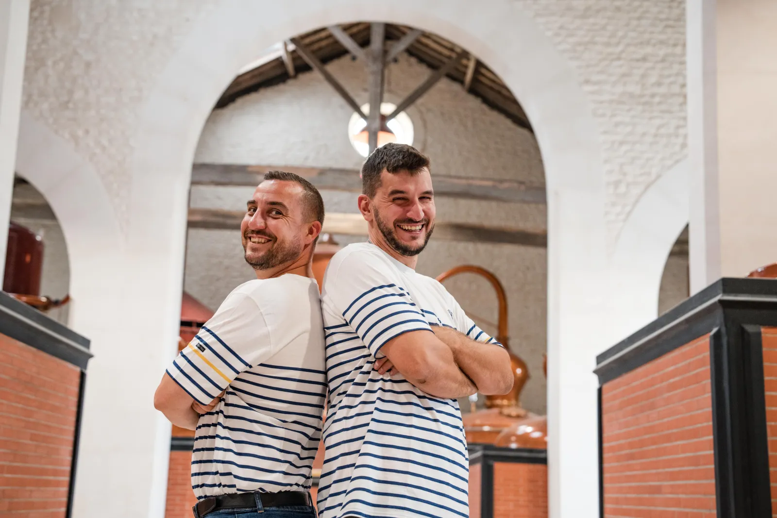 Two smiling men wearing blue and white striped shirts, standing back-to-back, posing in the white arched architecture of Citadelle Gin Distillery in Bordeaux.