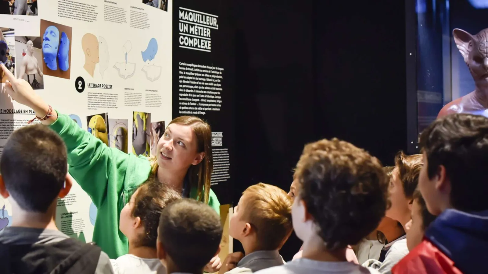 A facilitator in a green vest explains a makeup exhibition to a group of children at a science museum. Informational panels and head models are visible in the background.