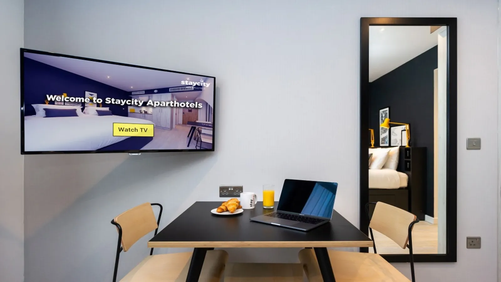 Staycity Aparthotel room in Bordeaux featuring a black work desk with beige chairs, laptop, breakfast items, and TV screen displaying a welcome message, with bedroom access visible in the background.