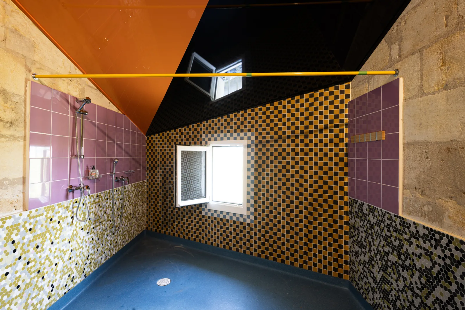 Shower room at the Joualles dormitory in Bordeaux featuring bold geometric tile patterns in yellow, black and multicolored designs, wall shower, window with shutters and vibrant orange ceiling