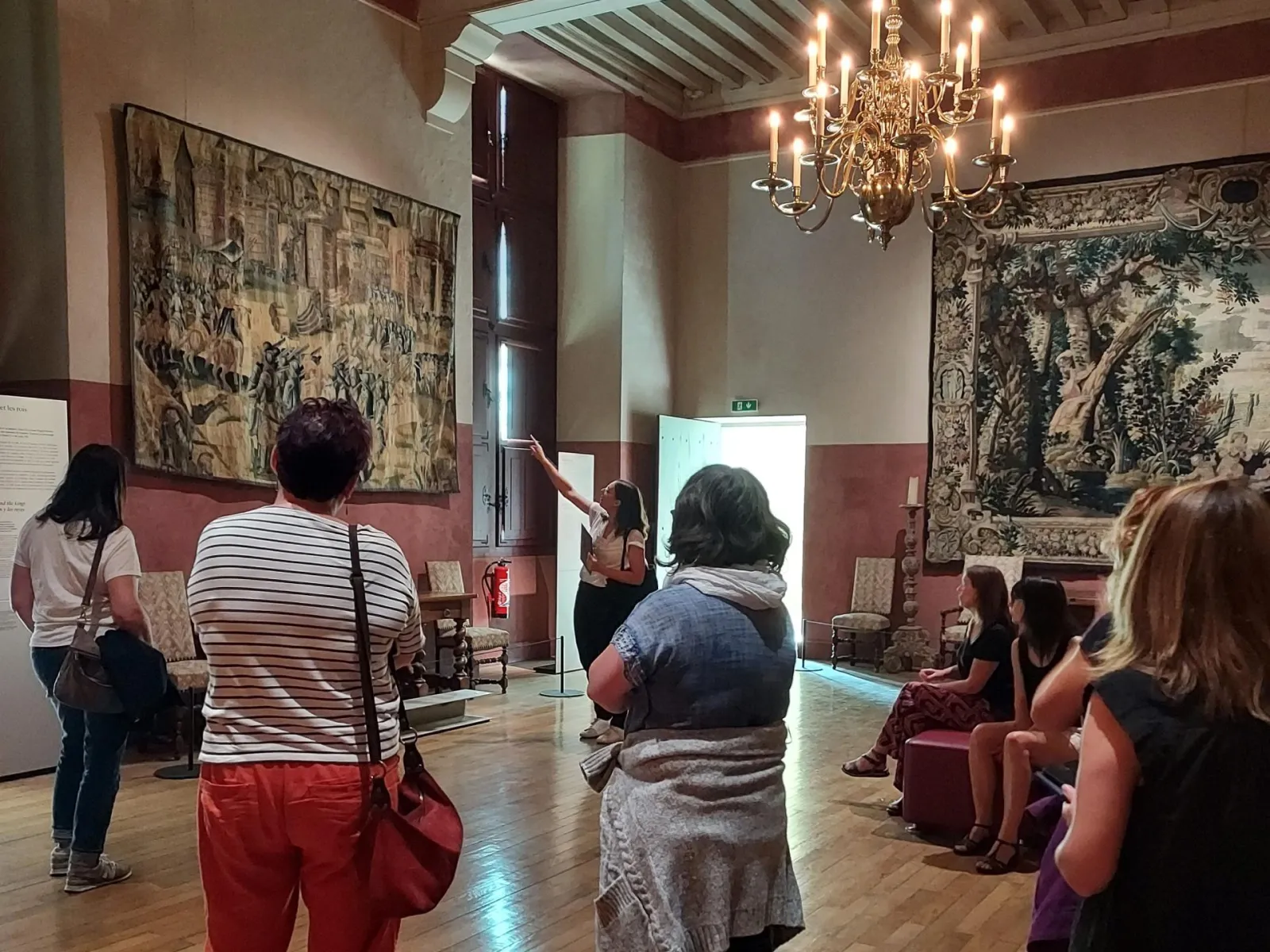 Guided tour in a room of Château ducal de Cadillac with visitors listening to a guide in front of historical tapestries and a crystal chandelier