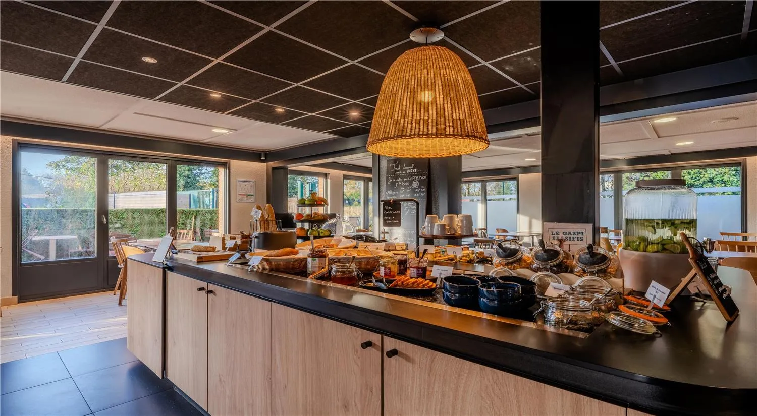 Breakfast room at Campanile Nature Bordeaux Lac featuring a service counter with pastries and breads, rattan pendant lamp, large windows overlooking the lake, and light wood furniture.