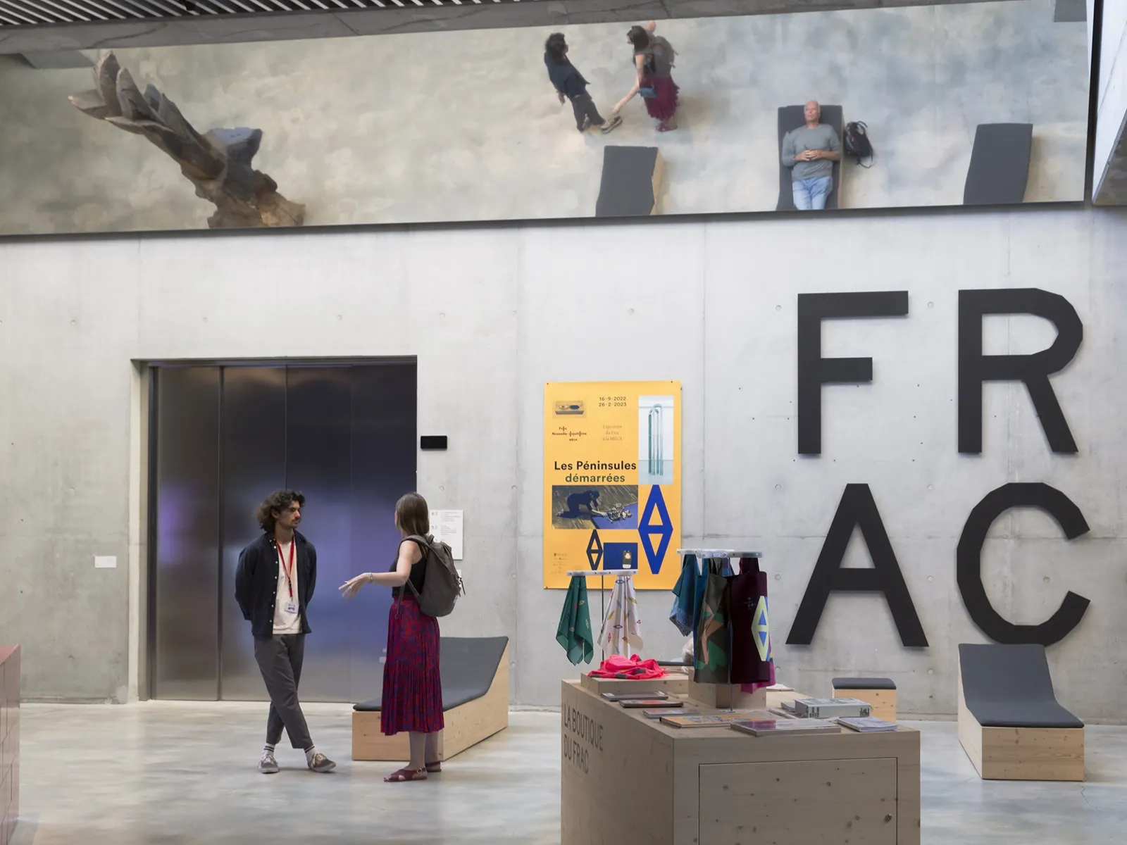 Interior of FRAC Nouvelle-Aquitaine MECA in Bordeaux: two people converse in a modern exhibition space with black panels, a yellow poster, and large 'FRAC' wall lettering. Hall view with visitors and sculpture in the background.